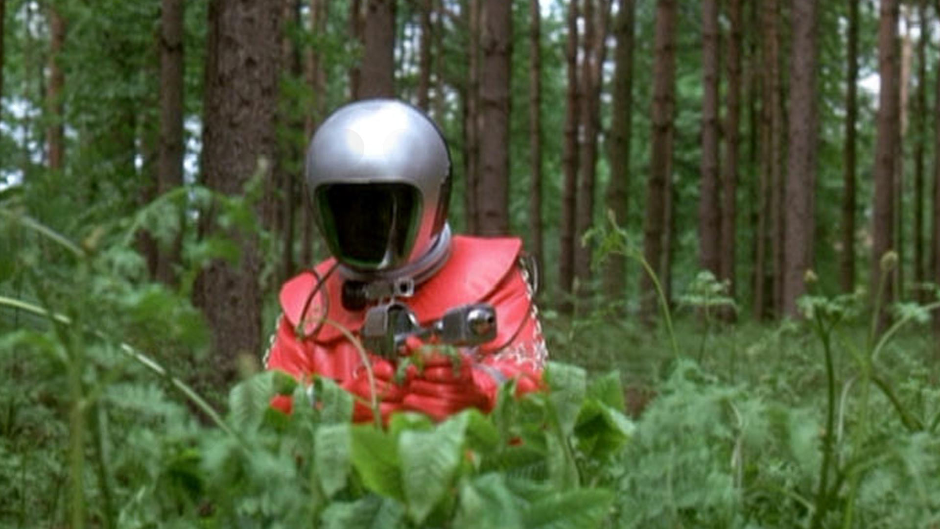 Scene from UFO (1970) - a red-suited alien figure with a mirrored helmet crouches in a pine forest